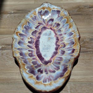 May include: An oval ceramic dish featuring a reactive glaze with a central white speckled area surrounded by concentric layers of deep purple, light blue, and warm tan tones. The edges of the dish are irregular and wavy, creating a natural form.