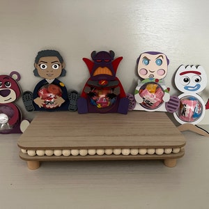 May include: Five Toy Story character themed candy holders. The holders are made of cardboard and feature a purple bear, a girl with brown hair, a purple and red villain, a blue and purple space ranger, and a white and red snowman. The holders are sitting on a wooden stand with a beaded trim.
