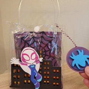 May include: Clear tote bag with a Spider-Gwen graphic. The bag features a cityscape cutout and a purple spiderweb design. A round tag with a blue spider is attached.