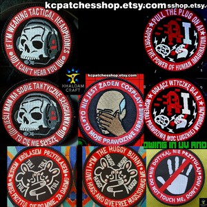 Może przedstawiać: Kolekcja haftowanych naszywek z różnymi wzorami i tekstami. Jedna naszywka przedstawia czaszkę w słuchawkach, inna mówi "PULL THE PLUG ON AI", a inne mają postacie z kreskówek i frazy. Naszywki są okrągłe i mają czerwone i białe akcenty.