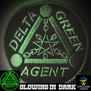 May include: A black and green embroidered patch with the words "Delta Green Agent" in a triangle shape. The patch glows in the dark.