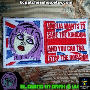 Może przedstawiać: Haftowana naszywka z fioletowowłosą kobietą i napisem "AMELIA WANTS TO SAVE THE KINGDOM AND YOU CAN TOO. STOP THE INVASION." Projekt zawiera brytyjską flagę w kolorach czerwonym, białym i niebieskim.