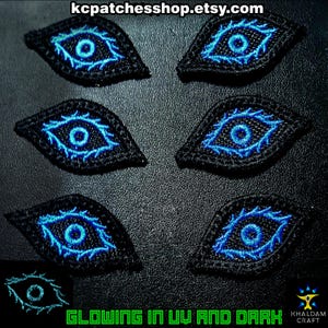 May include: Six black eye patches with blue glowing details. The patches have a detailed eye design with a central pupil and surrounding intricate lines. The text "GLOWING IN UV AND DARK" is visible at the bottom, along with a website address.