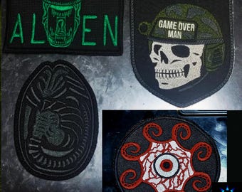 Op de afbeelding: Een verzameling geborduurde patches met verschillende ontwerpen. Eén patch toont het woord "ALIEN" met een groen wezenontwerp. Een andere toont een schedel met een helm met de tekst "GAME OVER MAN". Andere patches zijn een alien-ei en een cirkelvormig ontwerp met een oog.