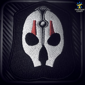 May include: A black embroidered patch with a white skull design. The skull has two red eyes and a white, stylized design on the forehead.