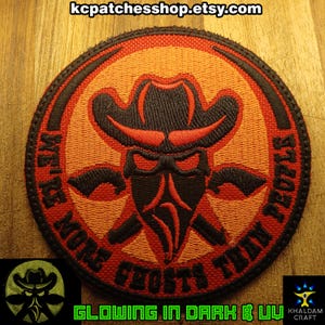Może przedstawiać: Okrągła naszywka z czarno-czerwonym wzorem przedstawiającym kapelusz kowbojski, skrzyżowane pistolety i napis "WE'RE MORE GHOSTS THAN PEOPLE". Naszywka świeci w ciemności i ma efekt UV.