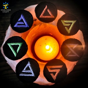 Może przedstawiać: Siedem czarnych łat z tkaniny z różnymi kolorowymi haftowanymi wzorami trójkątów, ułożonych wokół zapalonej świecy. Świeca znajduje się w szklanym świeczniku na białej powierzchni.