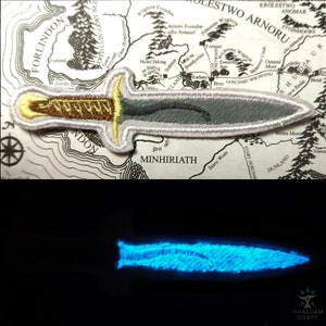 Könnte beinhalten: Ein grauer und goldener gestickter Aufnäher mit einem Dolch mit weißem Rand. Der Aufnäher befindet sich auf einer Karte von Mittelerde aus Der Herr der Ringe. Der Dolch leuchtet im zweiten Bild blau.