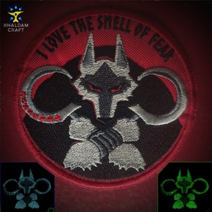 May include: A red circular embroidered patch featuring a white wolf with black eyes and a black mouth. The wolf is surrounded by two white circles with red accents. The text "I LOVE THE SMELL OF FEAR" is written in white around the edge of the patch.