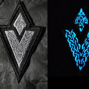 Może przedstawiać: Naszywka haftowana w kolorze czarnym i białym w kształcie litery V z mniejszym V wewnątrz. Naszywka świeci na niebiesko w ciemności.