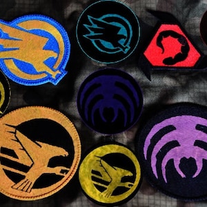 May include: A collection of eight embroidered patches featuring various designs. The patches are circular and feature a variety of colours, including black, white, yellow, blue, red, and purple. The designs include a bird with wings spread, a scorpion, and a spider.