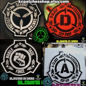 以下が含まれることがあります： 黒い背景に白いステッチで刺繍された4つのパッチ。パッチには、異なるテキストとシンボルが書かれたSCP財団のロゴが描かれています。パッチのテキストには、「確保、収容、保護」、「Dクラス職員」、「機動部隊」、「Aクラス職員」が含まれています。