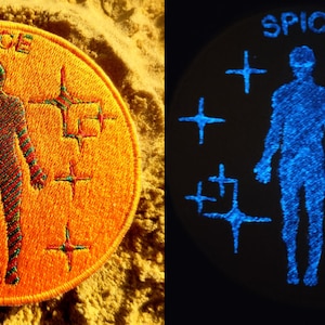 Może przedstawiać: Okrągła, pomarańczowa naszywka haftowana z napisem "SPICE" w białych literach. Naszywka przedstawia stylizowaną postać ludzką z niebieskim i czerwonym wzorem, otoczoną sześcioma białymi gwiazdami.