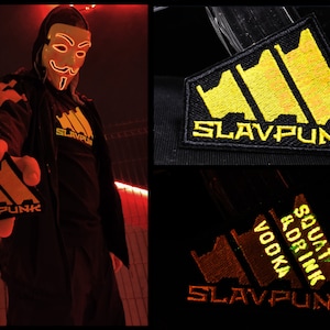 Könnte beinhalten: Eine Person trägt eine schwarze Kapuzenjacke und eine schwarze Maske mit gelbem Leuchten. Die Person hält ein gelbes und schwarzes Patch mit dem Text "SLAVPUNK" darauf. Das Patch ist ein stilisiertes Design aus drei vertikalen Balken.