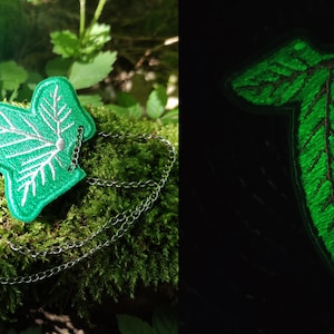 Puede incluir: Un collar con forma de hoja verde con una cadena plateada. La hoja brilla en la oscuridad.