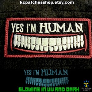 Może przedstawiać: Haftowana naszywka z napisem "YES I'M HUMAN" nad uśmiechniętym wzorem ust. Naszywka ma czarne tło z czerwoną obwódką. Poniżej wyświetlany jest tekst "YES I'M HUMAN" i świecący wzór ust, z napisem "GLOWING IN UV AND DARK" poniżej.