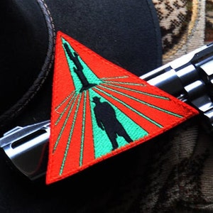 Op de afbeelding: Een rode driehoekige patch met een groen centrum en een zwart silhouet van een persoon die naar een lichtbron loopt. De patch heeft rode stiksels rond de randen en is geborduurd met witte lijnen.