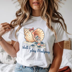 Pode incluir: T-shirt branca com um desenho de concha e peças de Mahjong. Um laço rosa adorna a concha. O texto "shell i play Mahjong" está escrito em azul por baixo. O design é uma versão lúdica do jogo clássico.