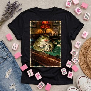 Könnte beinhalten: Ein schwarzes Kurzarm-T-Shirt mit einem rechteckigen Grafikdruck eines grünen Kröten, die auf einem grünen Filztisch sitzt. Die Kröte hält einen Mahjong-Stein mit chinesischen Schriftzeichen. Der Hintergrund zeigt eine Lampe und einen Holztisch.