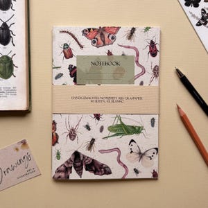 Könnte beinhalten: Ein Notizbuch mit beigem Einband, verziert mit Illustrationen von Insekten wie Schmetterlingen, Käfern und Würmern. Das Notizbuch ist mit einem beigen Band mit deutschem Text umwickelt. Ein Bleistift und ein Stift liegen neben dem Notizbuch. Das Notizbuch hat das Format A5.