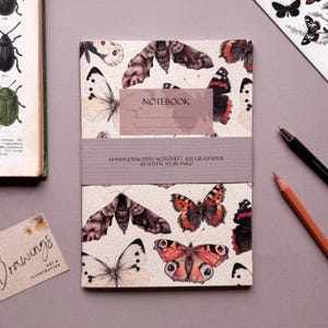 Könnte beinhalten: Ein Notizbuch mit einem Schmetterlingsmuster. Das Cover zeigt Illustrationen von Schmetterlingen in Braun-, Rot- und Weißtönen. Das Notizbuch ist mit einem mauvefarbenen Band mit dem Text "NOTEBOOK" und deutschem Text gebunden. Ein Bleistift und ein Stift sind sichtbar.