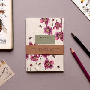 Op de afbeelding: Een notitieboek met een bloemenontwerp met paarse bloemen en bijen. De kaft heeft het woord "NOTEBOOK" op een groene rechthoek. Een bruine band wikkelt zich om het notitieboek met Duitse tekst. Een potlood en pen liggen naast het notitieboek.
