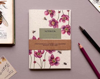 May include: A notebook with a floral design featuring purple flowers and bees. The cover has the word "NOTEBOOK" on a green rectangle. A brown band wraps around the notebook with German text. A pencil and pen are next to the notebook.