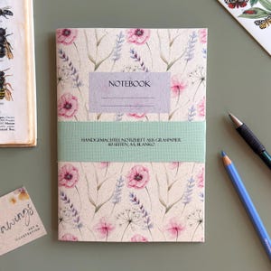 Könnte beinhalten: Ein rechteckiges Notizheft mit hellbeigem Einband und einem wiederkehrenden Muster aus rosa Mohnblumen und violetten Lavendelzweigen. Ein hellviolettes Etikett trägt die Aufschrift NOTEBOOK, und eine mintgrüne Papierbanderole zeigt den Text HANDGEMACHTES NOTIZHEFT AUS GRASPAPIER 40 SEITEN, A5, BLANKO.