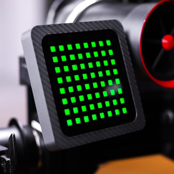 May include: A black and gray device with a green LED display. The device has a carbon fiber texture and is mounted on a black stand.