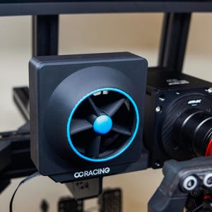 May include: A black fan with a blue accent ring and center, mounted on a racing simulator. The fan has the text "OCRACING" printed on the front. The simulator also includes a steering wheel and pedals.