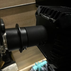 May include: Close-up of a black Fanatec racing wheel base and steering wheel assembly. The wheel base is rectangular with the brand name visible. The steering wheel is attached with a black cylindrical shaft and a silver ring.