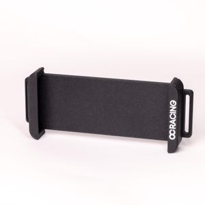 May include: A black phone holder with the text "OCRACING" printed in white. The holder has a rectangular design with two side clamps and a small loop on the side. The product is designed to hold a phone.