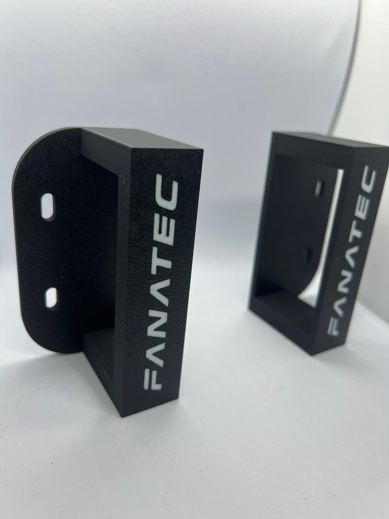 Puede incluir: Dos soportes de pared de pl&aacute;stico negro con la palabra "FANATEC" impresa en blanco en la parte delantera.