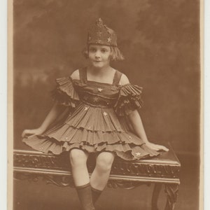 May include: A young girl wearing a crown with stars sits on a wooden bench. She is wearing a dark dress with ruffles and white stars. The girl is looking at the camera.