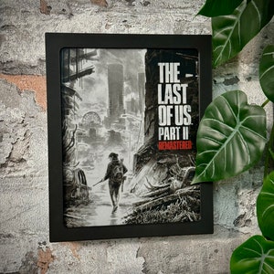 Könnte beinhalten: Ein schwarzer gerahmter Druck eines Videospiel-Covers für "The Last of Us Part II Remastered". Das Cover zeigt eine Frau in einer schwarzen Jacke und Jeans, die durch eine postapokalyptische Stadt geht.