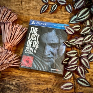 Könnte beinhalten: Ein Videospiel-Etui für The Last of Us Part II Special Edition für Playstation 4. Das Etui ist schwarz mit einem Nahaufnahmebild eines Gesichts. Das Etui hat einen roten USK 18-Aufkleber.