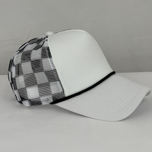 May include: White trucker cap featuring a black and white chequered mesh back and a white front panel. The cap has a black rope detail along the brim and a curved peak.