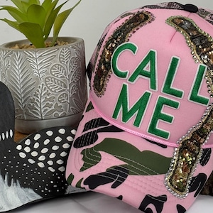 Puede incluir: Gorra de béisbol rosa camuflaje con el texto "CALL ME" en verde. La gorra presenta diseños de botellas con lentejuelas y una parte trasera de malla negra. Un somorgujo de madera y una planta en maceta están en el fondo.