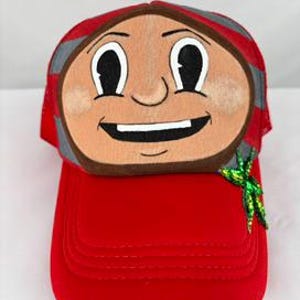 May include: A red trucker cap featuring a cartoon face patch on the front panel. The patch depicts a tan-skinned character with large black eyes, a wide smile, and rosy cheeks. A small, bright green sequined leaf accent is attached to the side.