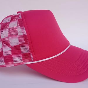 May include: A bright pink trucker cap featuring a white and pink chequered mesh back panel. The front of the cap is solid pink, with a white rope detail along the brim. The brim is curved and has stitched detailing.