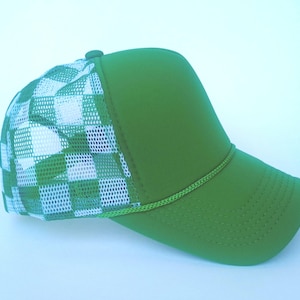 Op de afbeelding: Een groene truckerpet met een wit en groen geblokt mesh-achterpand. De pet heeft een effen groen voorpaneel en klep, met een bijpassend groen koorddetail langs de klep. Een klassieke stijl, geschikt voor vrijetijdskleding.
