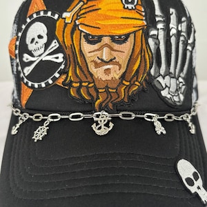 May include: Black trucker hat with a pirate design. It features a pirate face, skull and crossbones, and a skeleton hand. A silver chain with charms of anchors, a ship's wheel, and a skull is attached to the brim.