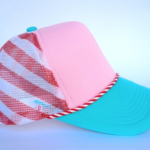 May include: A trucker hat with a pink crown, a turquoise brim, and a white mesh back with red stripes. The brim is trimmed with a red and white rope. A small turquoise button is on top.