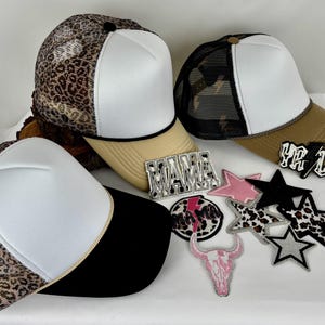 May include: A collection of trucker hats with white foam fronts, leopard print mesh or patterned fabric, and various colored brims. Patches with the words "MAMA" and "Y'ALL", stars, and a pink skull are also displayed.