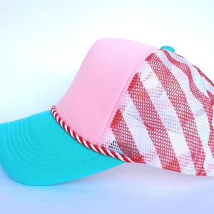 May include: A colorful trucker hat with a pink front panel, a turquoise brim, and a red and white striped mesh back. The brim is outlined with a red and white rope trim. A small blue button is on top.