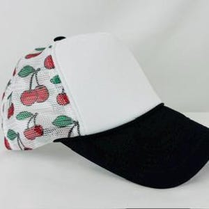 May include: A trucker hat with a white front panel and a black brim. The side panels are white mesh with a pattern of red cherries and green leaves. The top button is black.