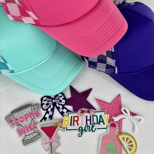 Könnte beinhalten: Mehrere farbenfrohe Trucker-Caps in Pink, Türkis und Lila mit karierten Mesh-Einsätzen. Bestickte Aufnäher mit Text und Designs wie Trophäe, Schleife, Martiniglas, Sterne und dem Schriftzug "Birthday Girl".