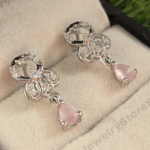 Peut inclure: Une paire de boucles d'oreilles pendantes argentées avec un motif floral et de petites pierres claires. Chaque boucle d'oreille comporte une pierre précieuse rose clair en forme de poire. Les boucles d'oreilles sont présentées dans un écrin noir avec un intérieur blanc.