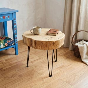 May include: A small, round wooden side table with black hairpin legs. The natural wood top has a live edge and is holding a mug, and a small notebook. The table is in a room with a blue table and a basket.