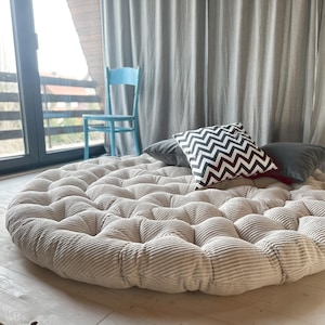 Puede incluir: Un cojín grande, redondo, de pana beige con un diseño acolchado. El cojín está sobre un suelo de madera y tiene dos almohadas encima. Una almohada es negra y la otra es negra y blanca con un patrón de chevron.